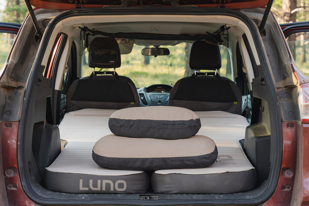 Air mattresses and two Packable Camp Pillows by Luno are set up in the back of a car with its rear doors open, designed for car camping or road trips. The car seats are folded forward, creating a flat sleeping area perfect for cozy nights on the road. This setup is ideal for anyone seeking portable comfort and practical gear from Luno during their adventures.