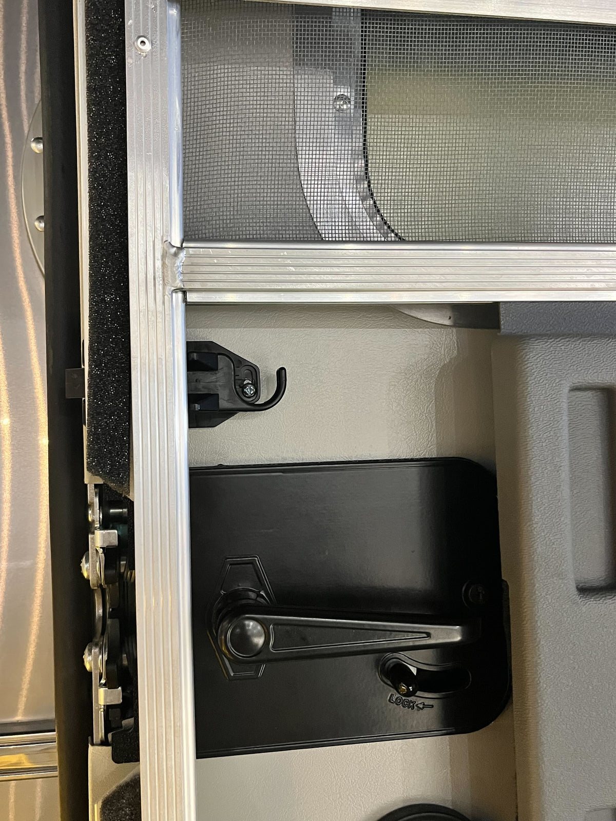 Close-up of a metal window frame with a screen, featuring a black handle and the Airstream Bullet Latch for Screen Door (Part #381454) with locking mechanism on a gray panel—perfect as a reliable replacement part for your Airstream or similar vehicle.