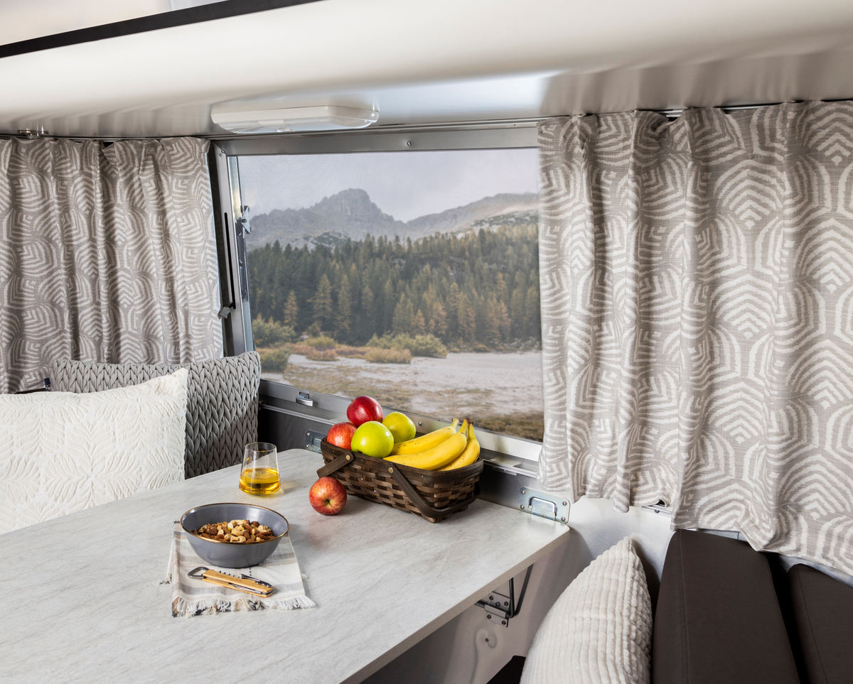 A cozy RV dining area featuring Airstream Printed Custom Curtains for Pottery Barn Travel Trailers, with a bowl of mixed nuts, a glass of juice, and a basket of fruit on the table. Through the window, mountains and a forested landscape are visible.