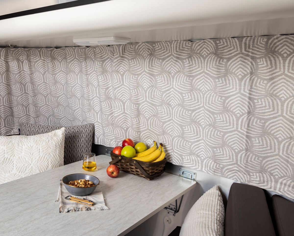 A dining table inside an Airstream Bambi camper is set with a drink, a bowl of nuts, and a basket of assorted fruits including bananas, apples, and lemons. Airstream Printed Custom Curtains for Bambi Travel Trailers and patterned cushions add charm to the cozy space.