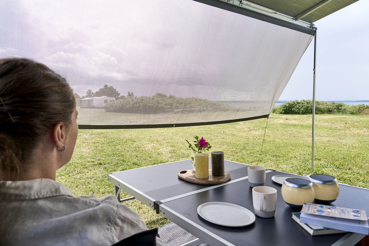 Relaxing at a table beneath the Shade Screen for Airstream Rangeline, a person enjoys views of a grassy field while cups, bowls, a book, and a tray with a drink, flower, and condiments rest on the tabletop. The shade screen provides sun protection and allows for an inviting outdoor experience.