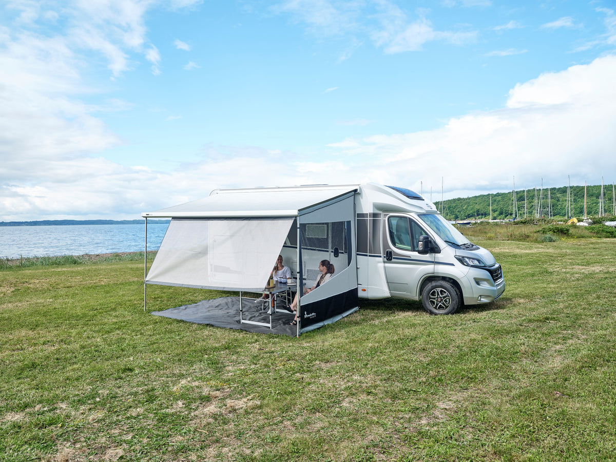 A camper van is parked on grass near a lake. Two people sit at a table under the Shade Screen for Airstream Rangeline, enjoying the outdoors with excellent sun protection. Sailboats and hills are visible in the background under a partly cloudy sky.