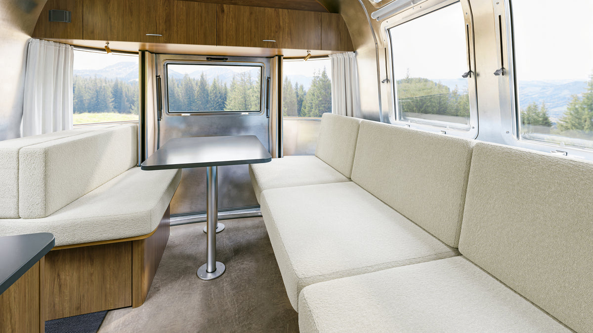 Interior of a camper featuring Airstream Custom Replacement Cushion Covers for Trade Wind Travel Trailers in a cream color on the seating, complemented by a small black table, wood-paneled cabinets, and large windows with views of trees and mountains outside.