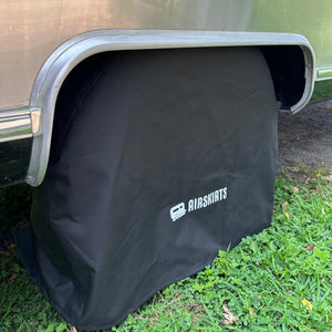 A black wheel cover from the Airstream Inflatable Skirt Kits by AirSkirts is positioned over a trailer’s wheel, partially covering the tire and rim, and rests on grass beside the vehicle to complement your RV skirting setup.