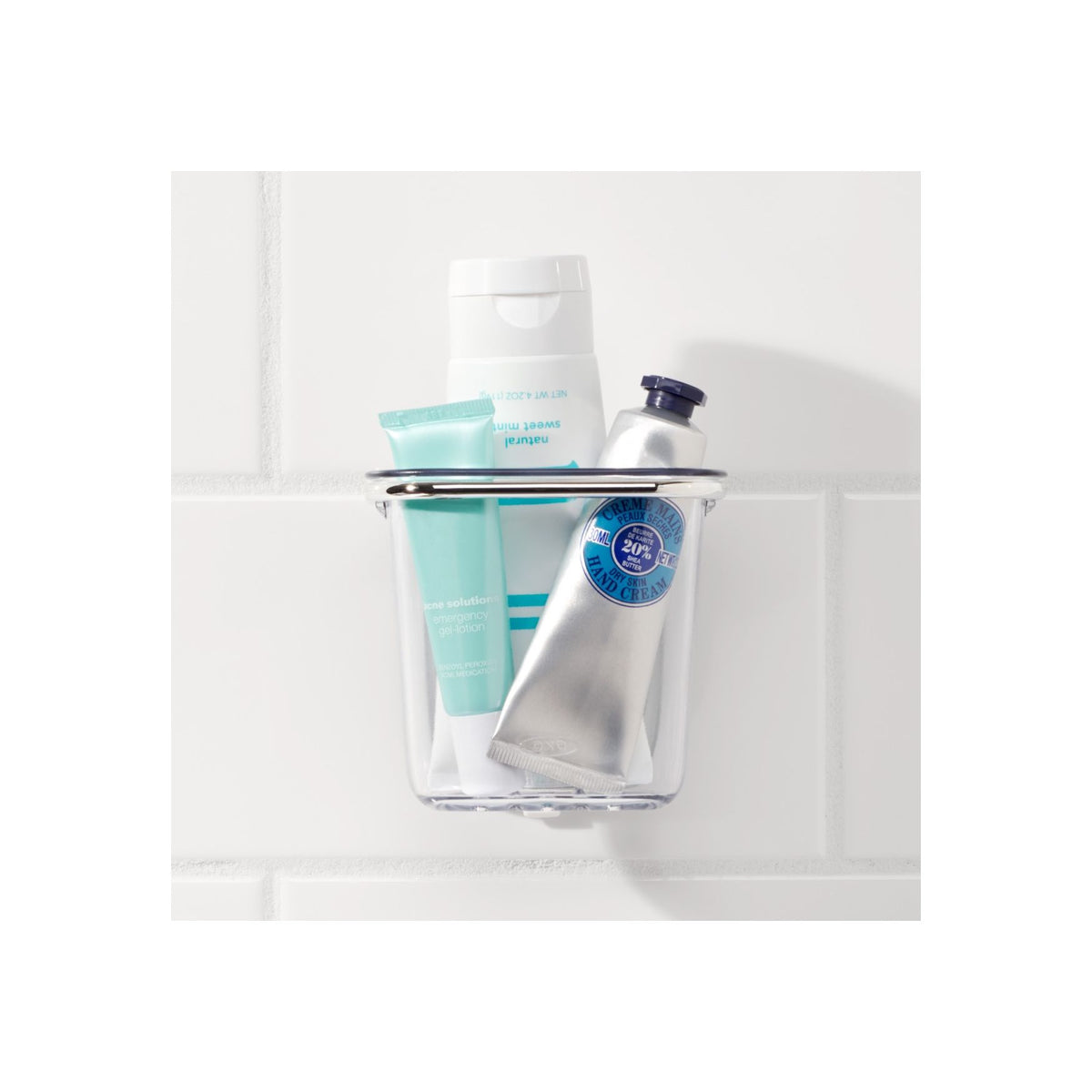 A clear wall-mounted Suction Basket by OXO, ideal as a storage solution for any space—from modern homes to an Airstream bathroom—holds three bath or skincare products: a white bottle, a turquoise tube, and a silver tube, all set against a white tiled background.