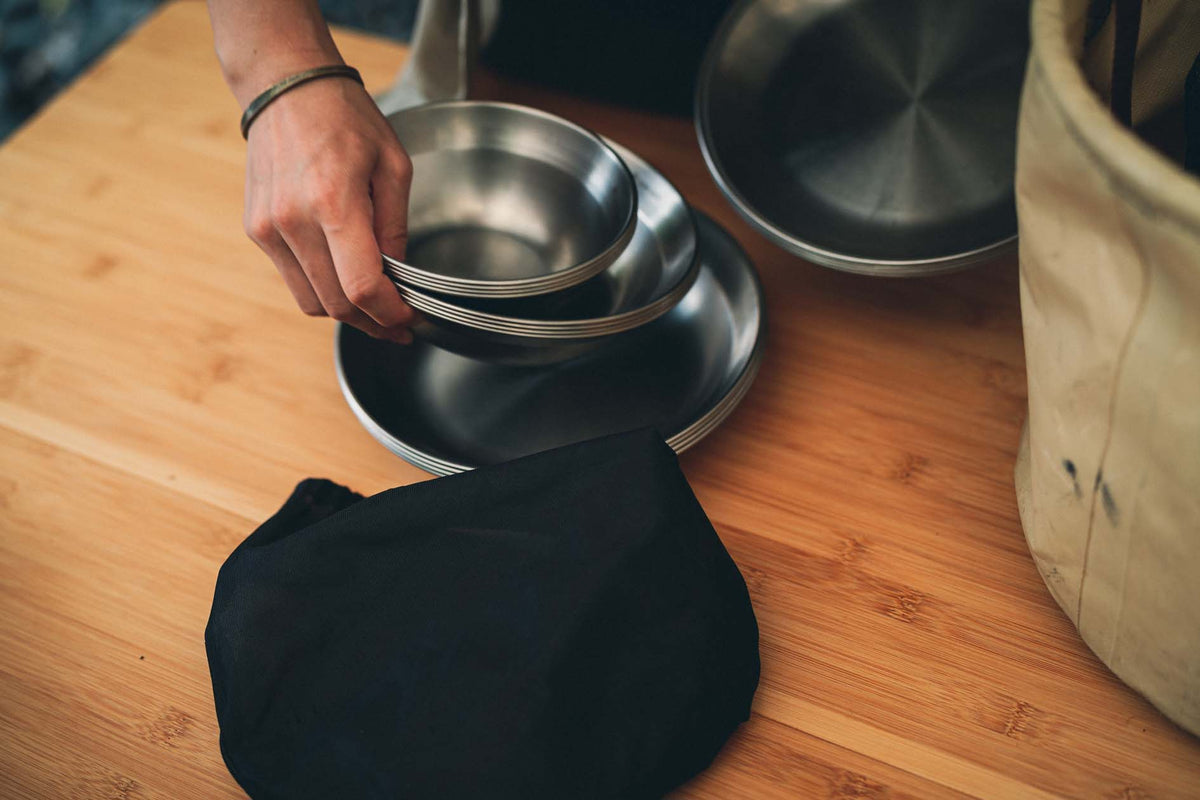 A persons hand is stacking metal bowls and plates on a wooden surface, with a black cloth bag and a beige bag nearby.