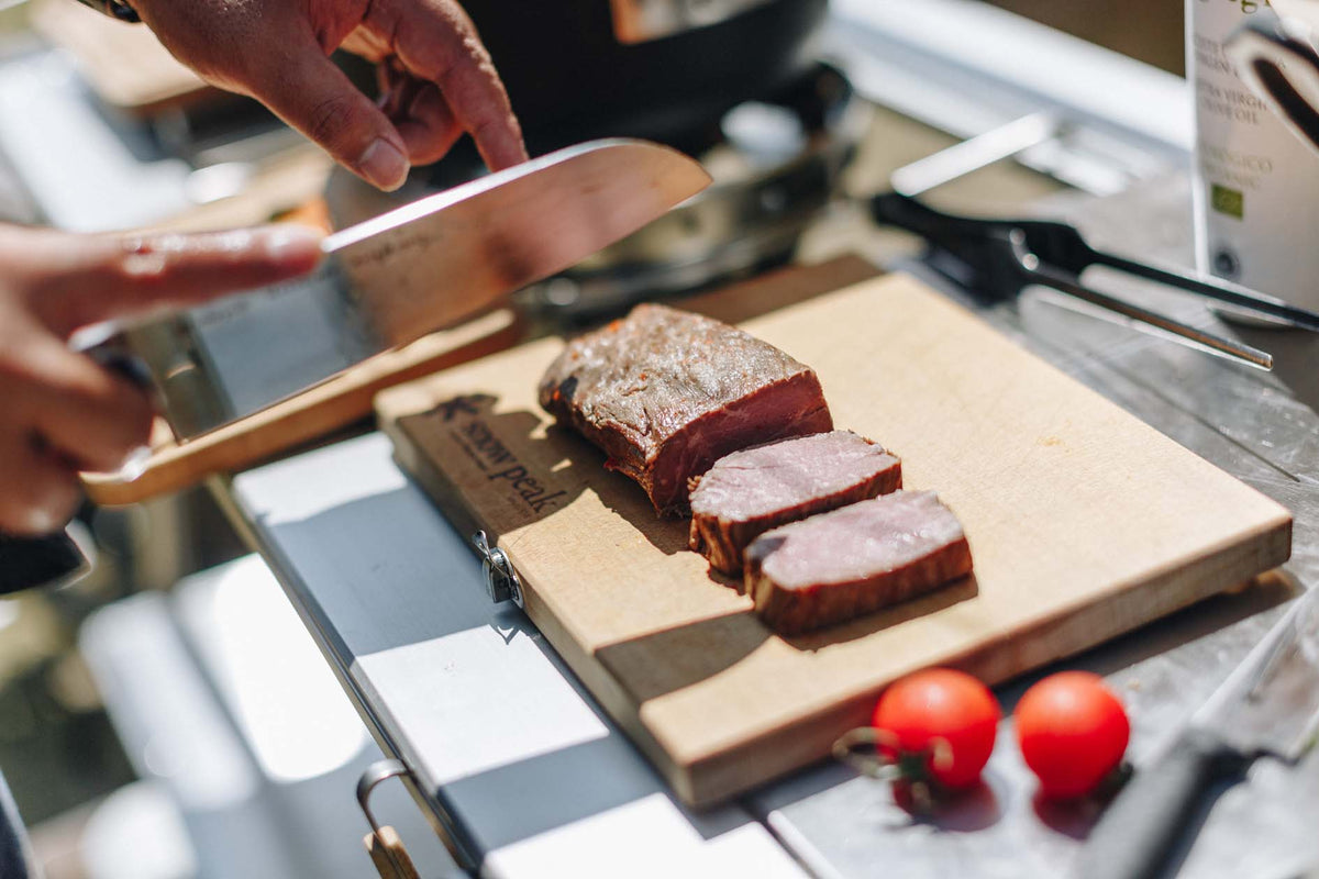 A person slices cooked steak on a wooden cutting board beside two cherry tomatoes and kitchen utensils on a countertop.