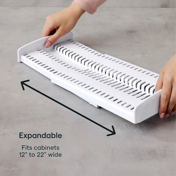 A person expands a white plastic organizer rack on a gray surface. Text reads: Expandable. Fits cabinets 12 to 22 inches wide. Black arrows indicate the direction of expansion, demonstrating the adjustable Airstream StoreMore Roof Locker Dish Rack by YouCopia—ideal for creating customized storage solutions in your kitchen.