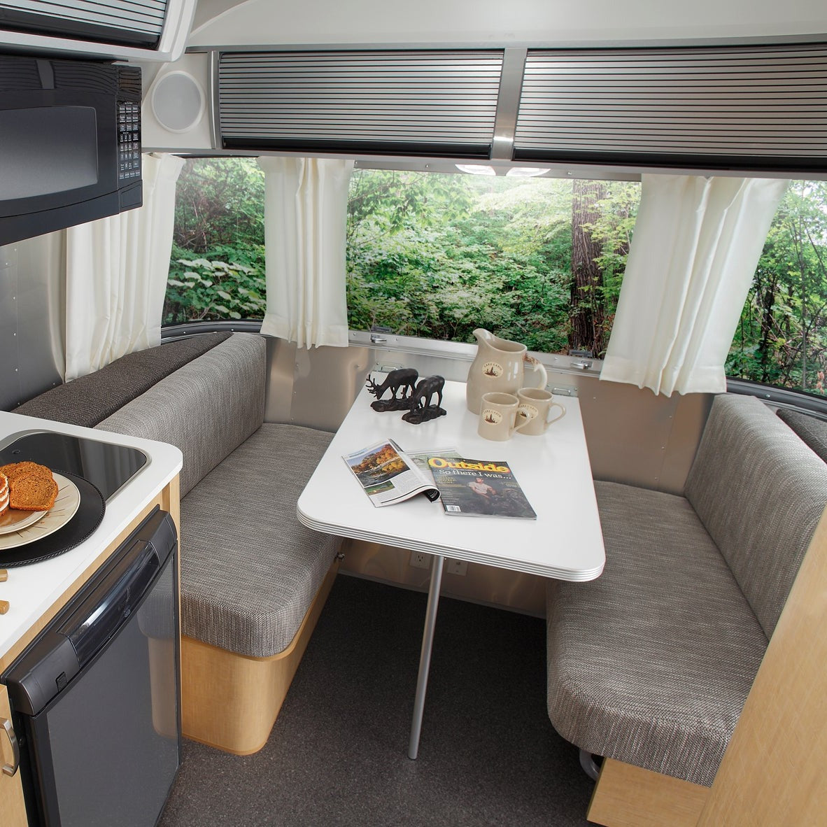 Cozy RV dining area featuring gray cushioned benches and a white table displaying magazines, mugs, and a bear figurine, with a large window dressed in Airstream Solid Custom Curtains for Sport Travel Trailers that frames views of lush greenery outside. Part of the small kitchen area is also visible.