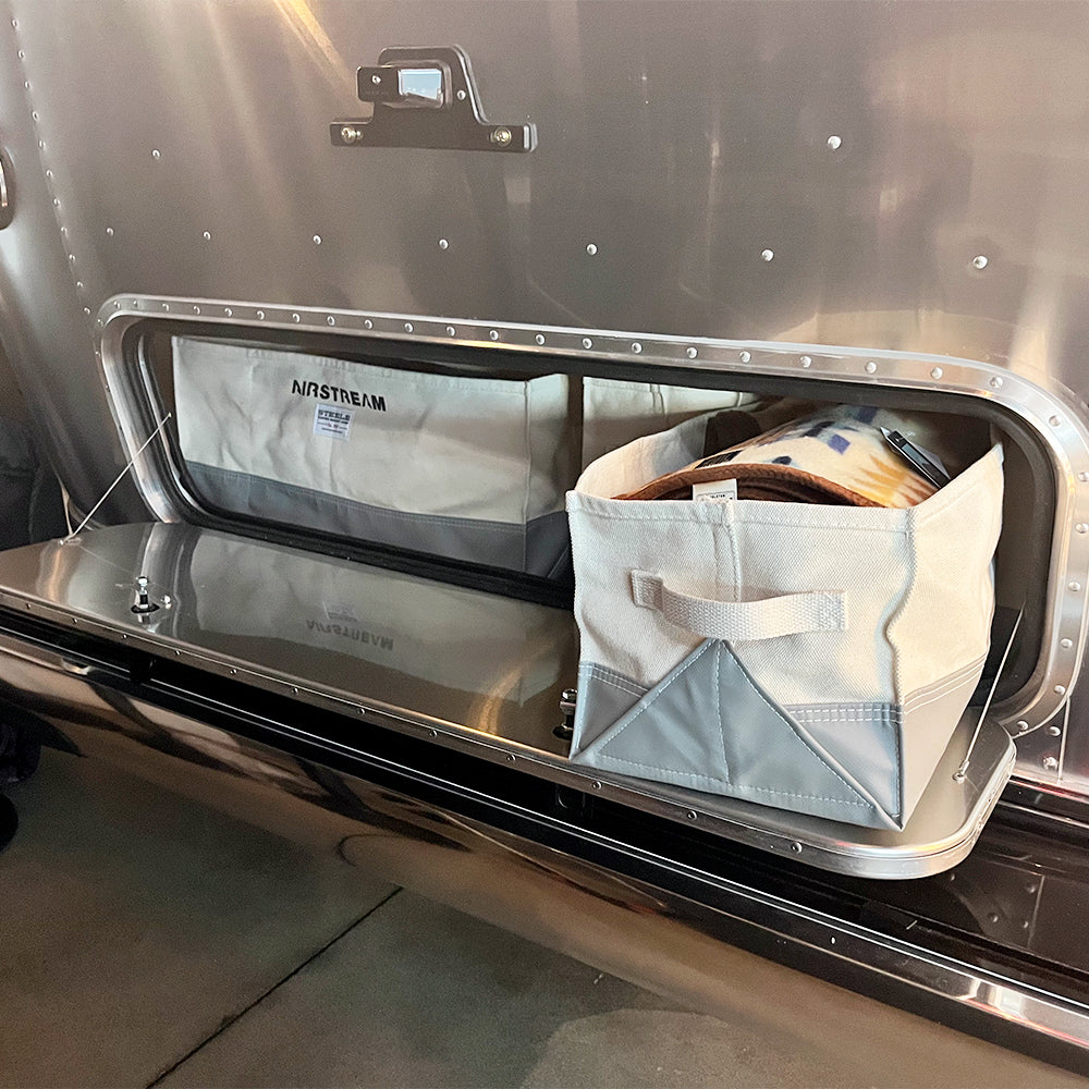 An open crossbed storage compartment on a shiny trailer reveals an Airstream x Steele Canvas Crossbed Storage Bin containing a folded blanket, alongside a bag marked Airstream. The compartment door is lowered, exposing metallic surfaces and prominent rivets.