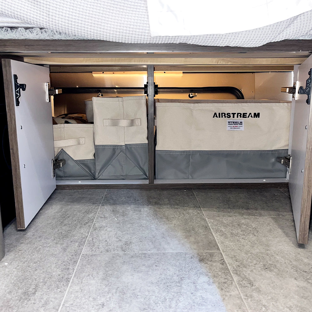 Open cabinet doors reveal organized Airstream x Steele Canvas Crossbed Storage Bins and additional crossbed storage beneath a bed, with gray tile flooring and a bed frame visible above the neatly arranged custom bins.