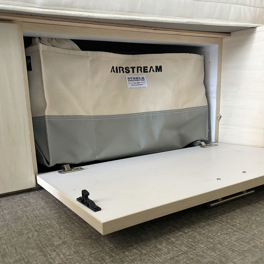 An Airstream x Steele Canvas Twin Bed Storage Bin is stored inside an open white twin bed floor cabinet with a fold-down door, revealing gray flooring and light wood paneling.