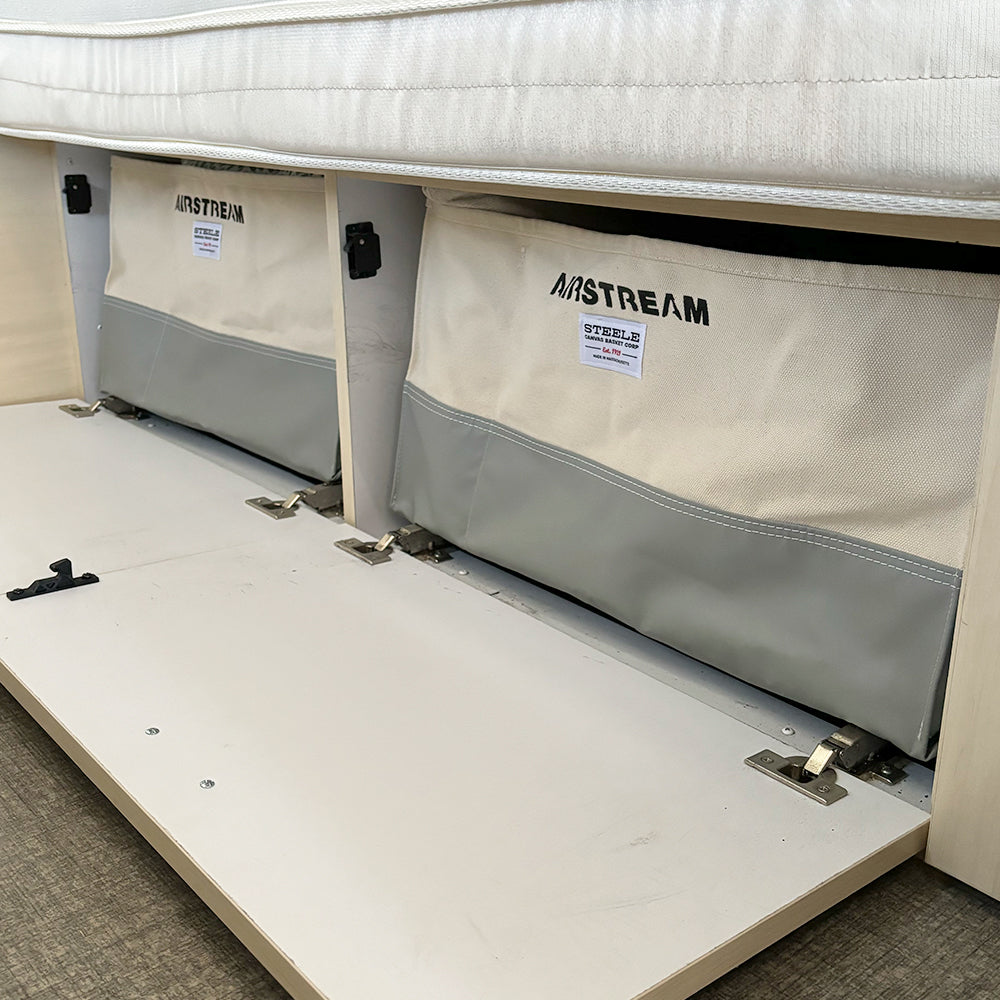 Open under-bed storage compartments reveal two Airstream x Steele Canvas Twin Bed Storage Bins, featuring heavy-duty canvas construction with gray bottoms and white tops, set in a light wood frame beneath the mattress.