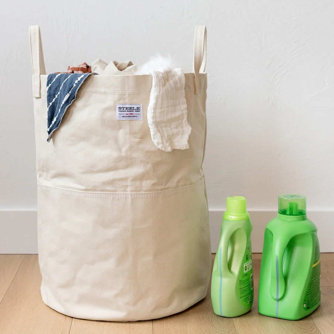 A Tall Canvas Storage Bin in beige, filled with towels and clothes, sits on a wooden floor next to two green bottles of laundry detergent—ideal for Airstream storage or camping essentials. A white wall serves as the backdrop.