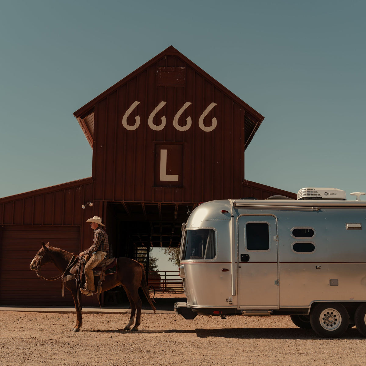 A person on horseback is next to an Airstream Stetson + 6666 Special Edition trailer in front of a large red barn marked with 6666 and a white L under a clear blue sky.