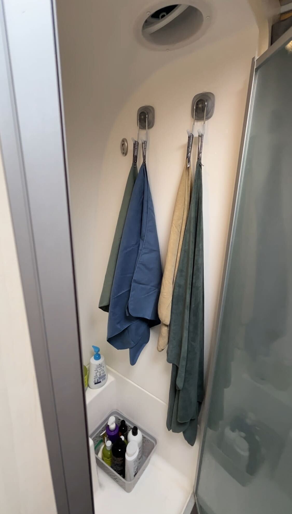 Two towels hang on StrongHold™ Suction Hooks by OXO in a compact shower. Bottles of toiletries, including soap and shampoo, are organized in a small bathroom organizer with a non-slip grip on the shower floor. A frosted glass shower door is partially visible.