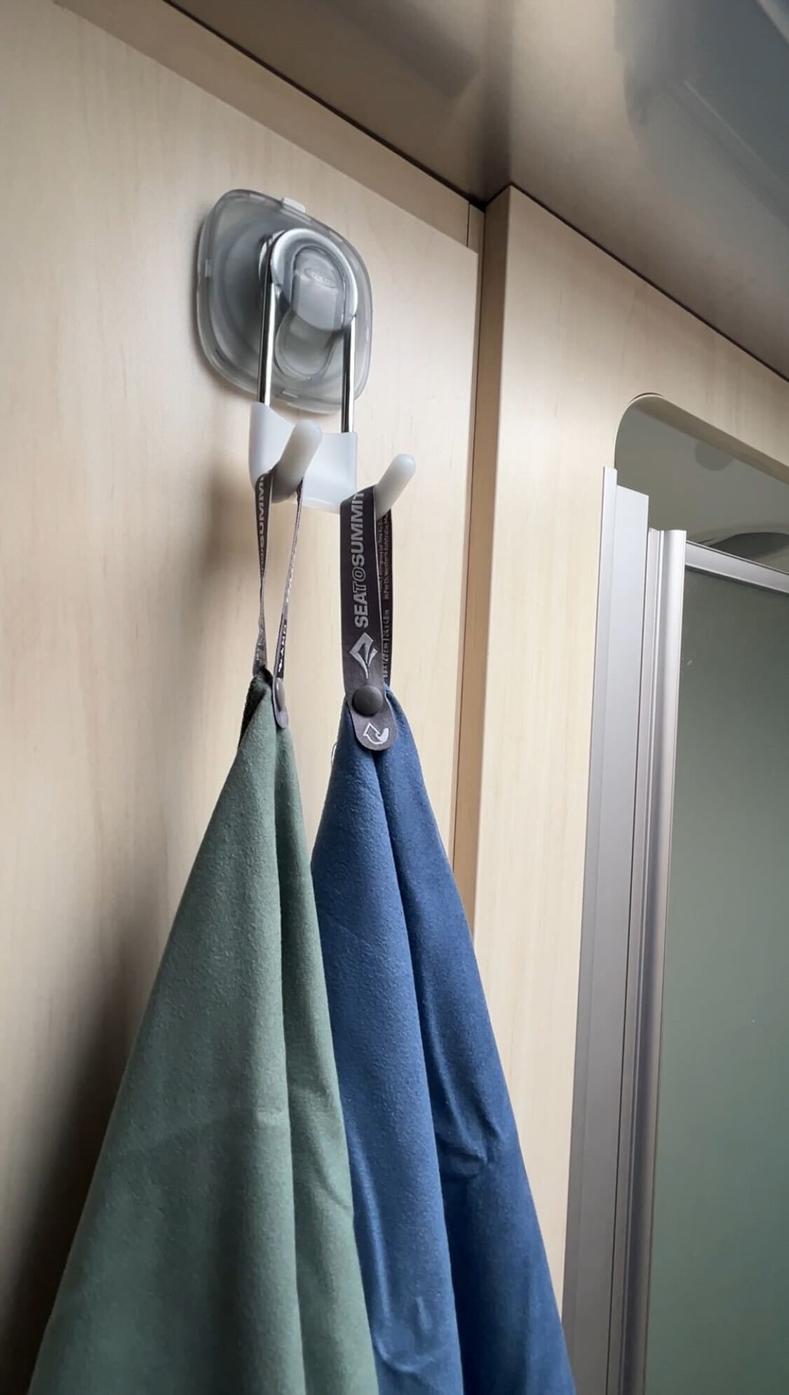 Two towels, one green and one blue, hang on a white StrongHold™ Suction Hook by OXO with a non-slip grip attached to a light wood wall. A frosted glass shower door is partially visible on the right.