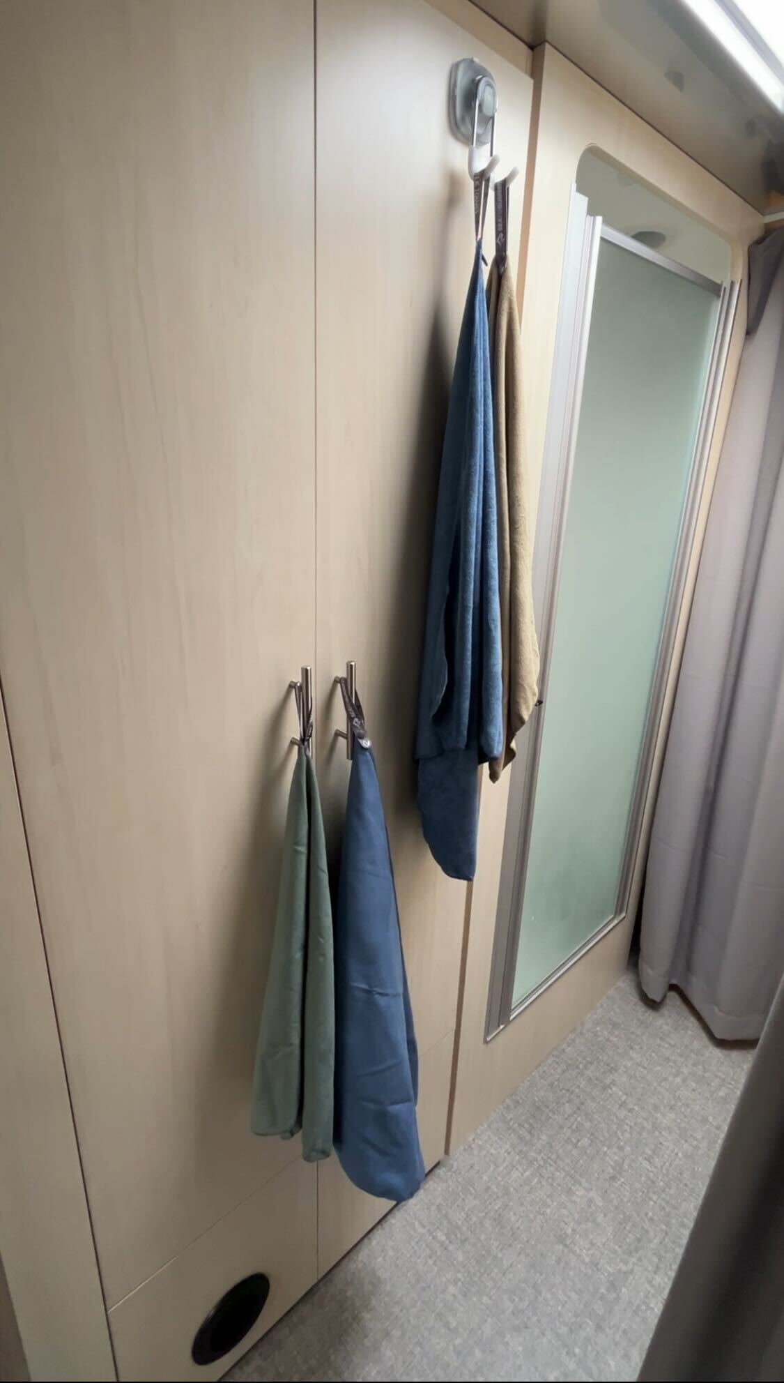 Four towels in blue, green, tan, and dark blue hang from StrongHold™ Suction Hooks by OXO attached to a light wood wall near a frosted glass shower door in a modern bathroom—creating an effortless organizer with reliable non-slip grip.