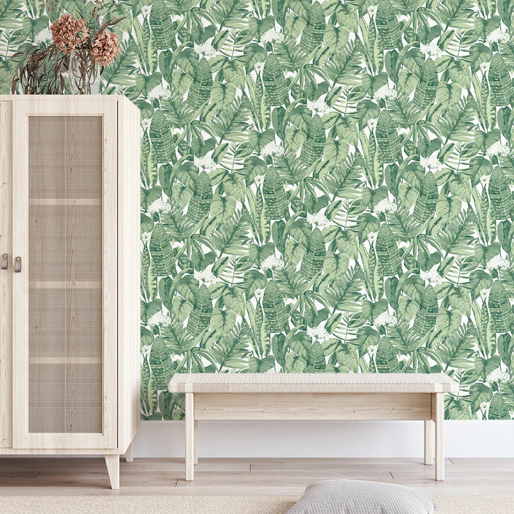 A room with a light wood cabinet, a matching bench, and walls decorated with Tropical Peel + Stick Removable Wallpaper featuring a green leafy pattern. Dried flowers in a vase sit on the cabinet. The floor is light wood with a beige textured rug.