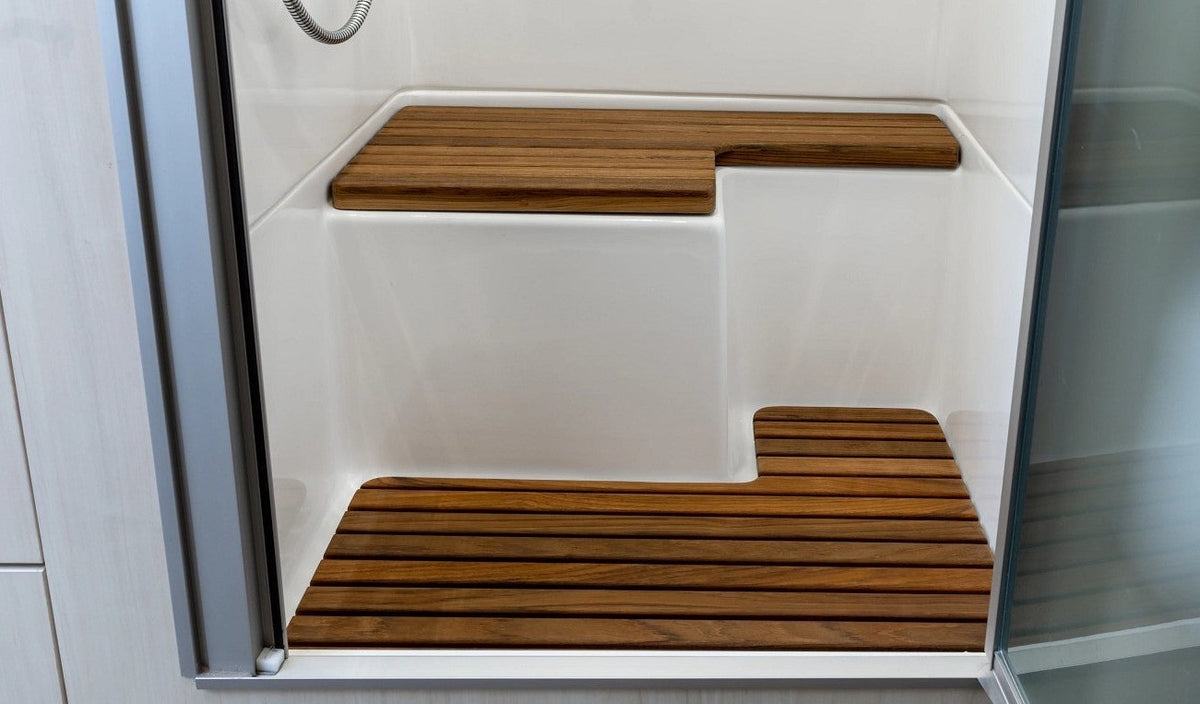 Shower stall with a built-in white seat and an Airstream Teak Shower Bench for International Travel Trailers placed on a wooden slatted floor mat, viewed through a partially open glass door.