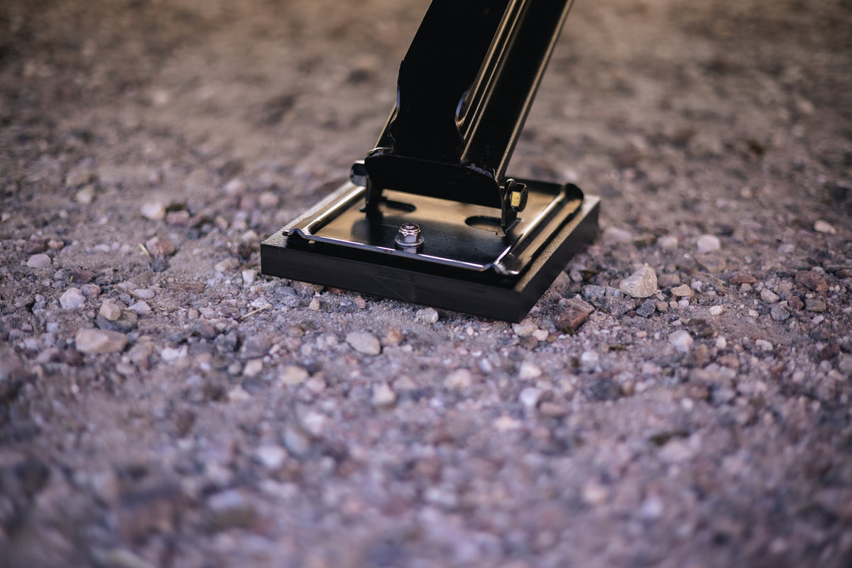 A close-up of a metal support plate resting on gravel, attached to a black angled metal bar, with a visible bolt securing the plate to the ground.