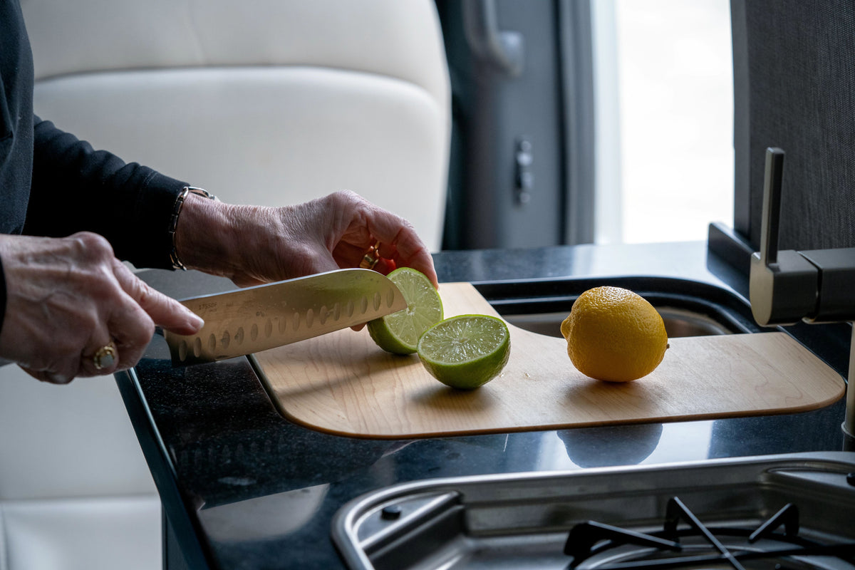 A person slices a lime on the Airstream Custom Sink Cutting Board for Interstate 24GT/GTX next to a whole lemon in a modern kitchen, with only their hands visible near the sink and stove.