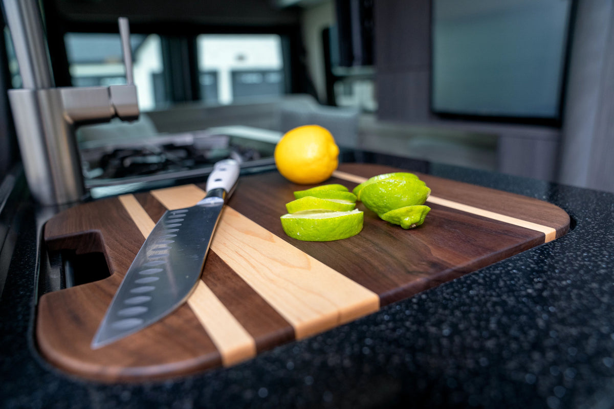 An Airstream Custom Sink Cutting Board for Interstate 24GT/GTX, featuring a stylish striped wood design, rests in a modern kitchen setting with a knife beside it, accompanied by a halved lime, three lime wedges, and a whole lemon.