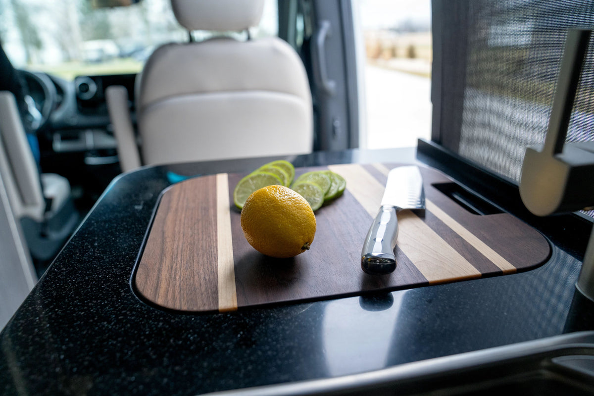 A lemon and sliced lime rest on an Airstream Custom Sink Cutting Board for Interstate 24GT/GTX with a knife, set on a black counter inside a van with the driver’s seat visible in the background.