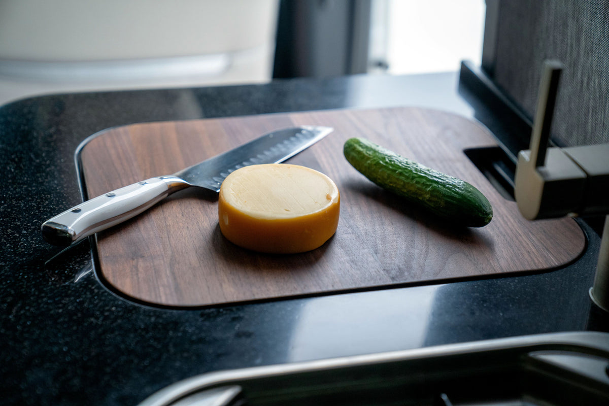 A knife, a round block of yellow cheese, and a cucumber sit on an Airstream Custom Sink Cutting Board for Interstate 24GT/GTX atop a dark kitchen counter.