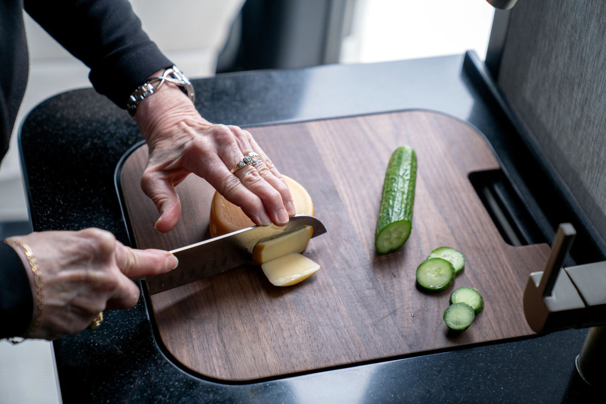 A person slices a round block of cheese on an Airstream Custom Sink Cutting Board for Interstate 24GT/GTX, placed on a wooden counter, with both sliced and whole cucumbers neatly arranged on the board.