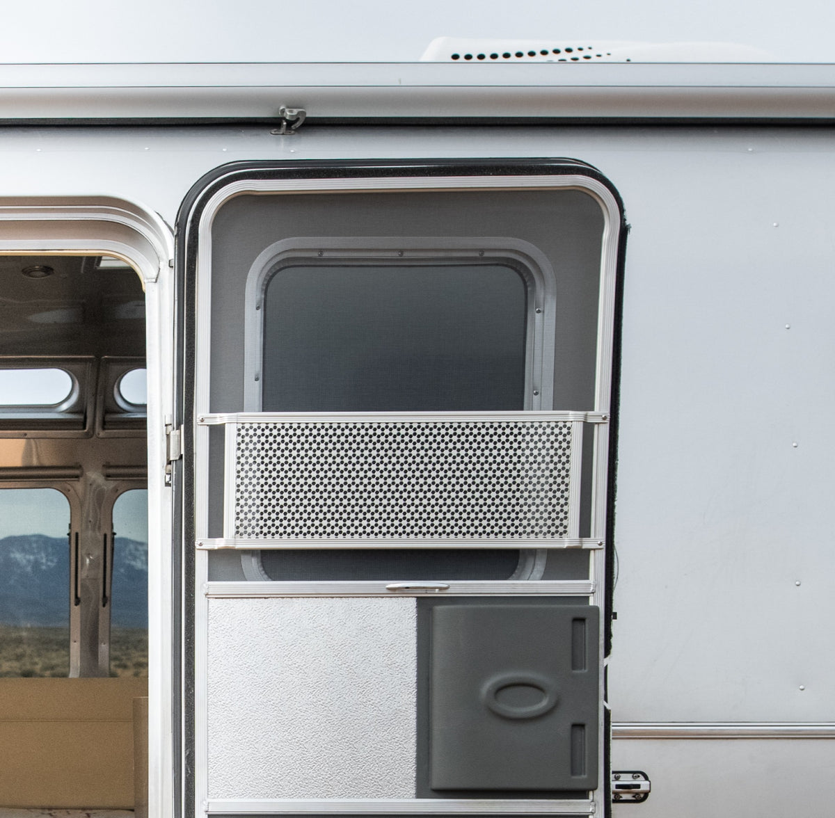 A close-up of an open silver camper door with a mesh screen, featuring the Airstream Screen Door Close Out (Part #203287-02). Mountains and plains are visible through the open doorway in the background.