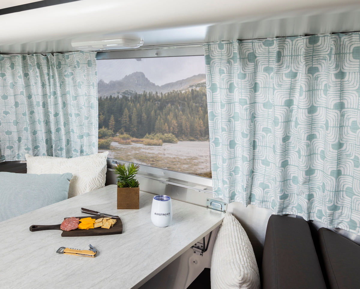 Inside the cozy RV dining area of an Airstream Bambi travel trailer, cushioned seats surround a table set with cheese, crackers, and salami. Charming Airstream Printed Custom Curtains for Bambi Travel Trailers hang at the windows, complementing the space alongside a potted plant and framing a scenic view of mountains and trees outside.