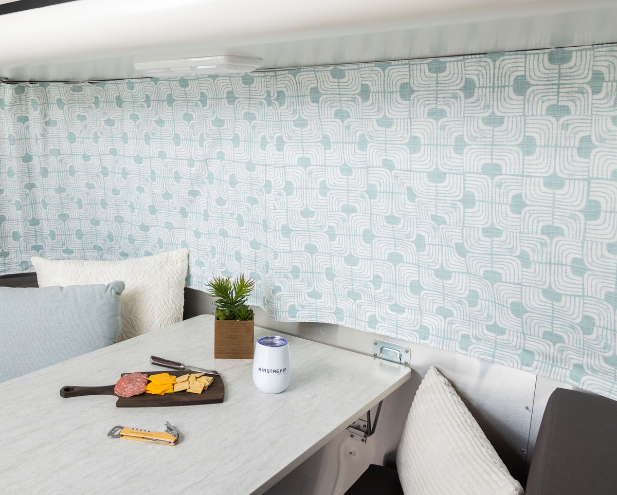A marble table with cheese, crackers, salami, a beverage can, a plant, and pillows is set inside a room featuring blue and white patterned Airstream Printed Custom Curtains for Bambi Travel Trailers.
