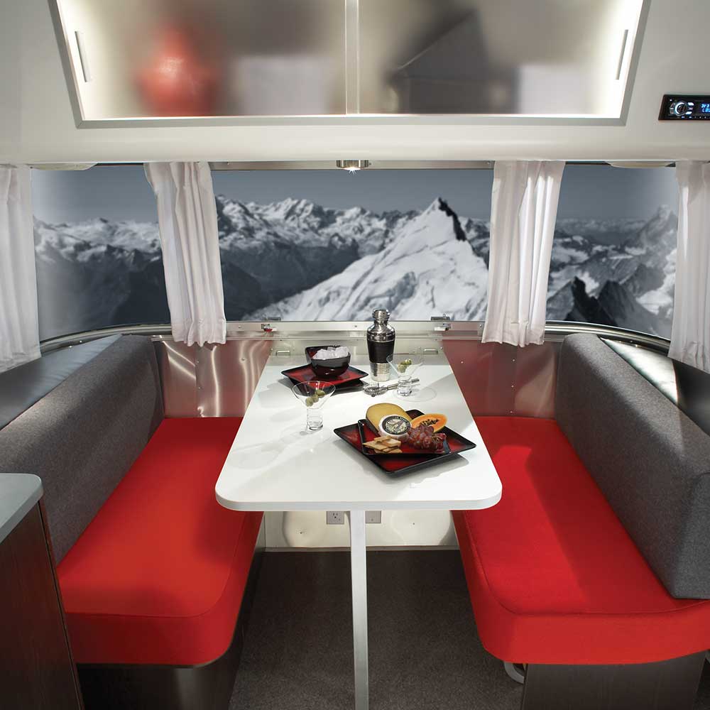 A modern RV dining area with red seats and a white table set with gourmet food and drinks, featuring large windows that reveal a snowy mountain landscape outside—elegantly framed by Airstream Solid Custom Curtains for Victorinox Travel Trailers, which offer both style and privacy.