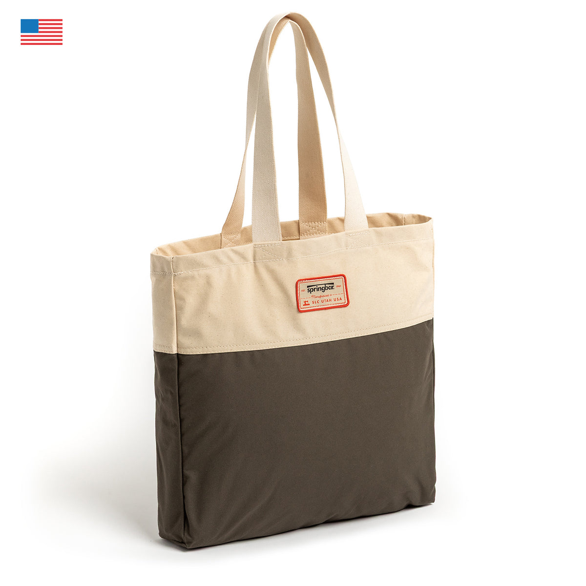 The Camp Tote by Springbar features a two-tone design with a beige top, dark olive bottom, and cream handles, accented by a rectangular red-bordered label on the front. Expertly crafted from durable Sunforger Cotton Duck Canvas, the bag stands upright on a white background, with a small American flag displayed in the top left corner.
