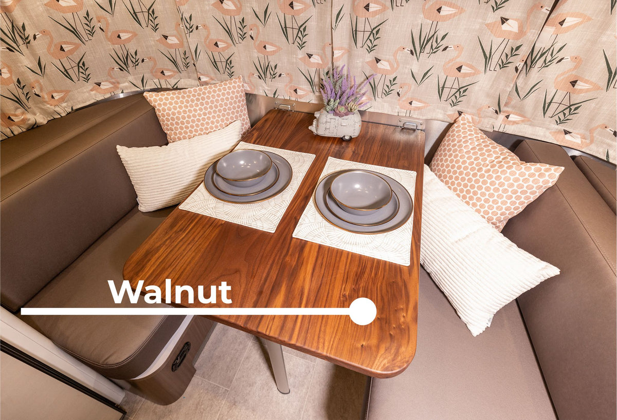 An Airstream Wood Dinette Table Upgrade for Flying Cloud Travel Trailers, featuring a walnut finish and solid wood construction, is set with plates and bowls, surrounded by brown cushioned seats and decorative pillows. Floral curtains frame the space, while a small lavender centerpiece adds a touch of charm.