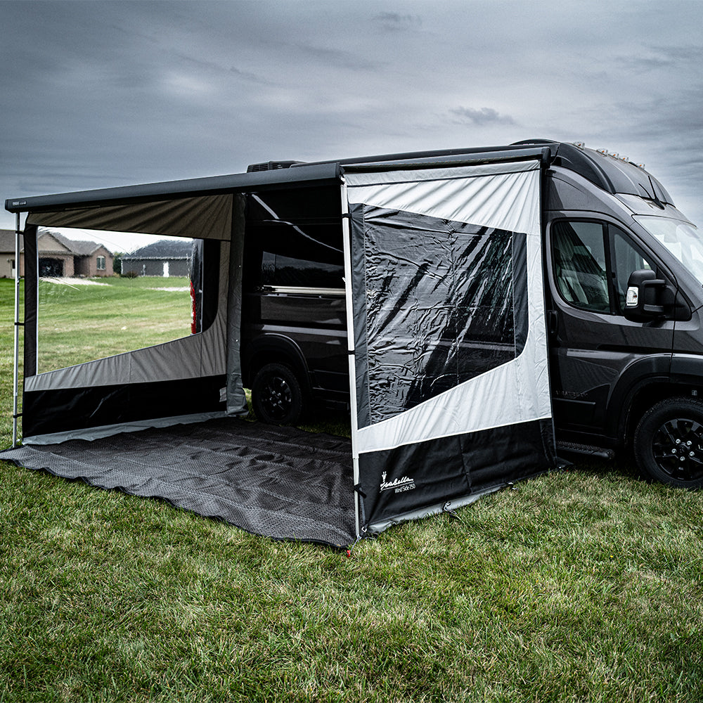 A black Airstream Rangeline camper van, featuring Wind Side Panels and an extended sun canopy, creates a covered outdoor space on the grass beneath a cloudy sky.