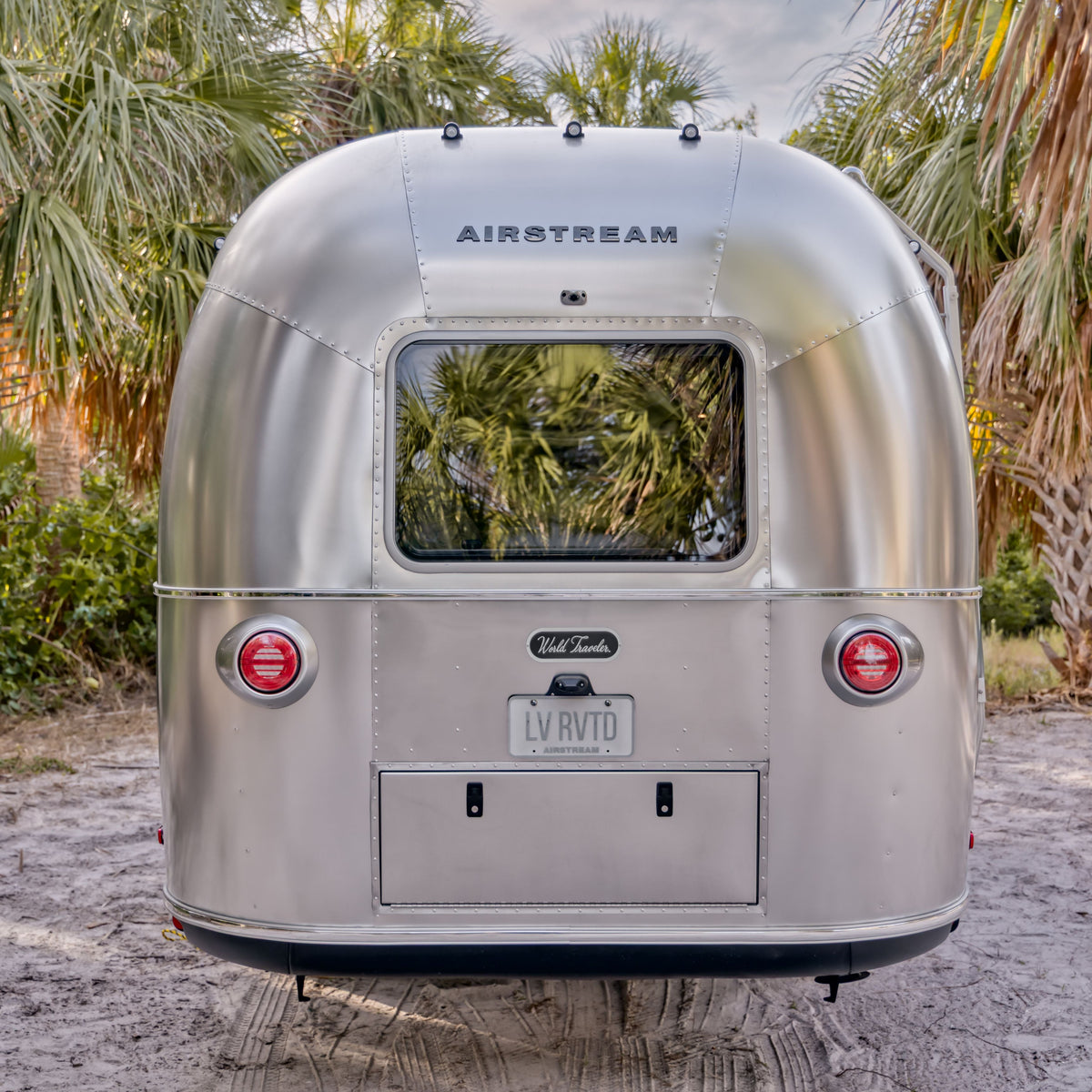 A silver Airstream travel trailer, reminiscent of those featured in the “Airstream Owner Manuals: World Traveler Travel Trailers,” is parked on sandy ground and encircled by palm trees. With its rear window, taillights, and license plate reading LV RVTD, this scene perfectly captures the spirit that inspires enthusiasts to explore owner manuals or imagine life on the open road.
