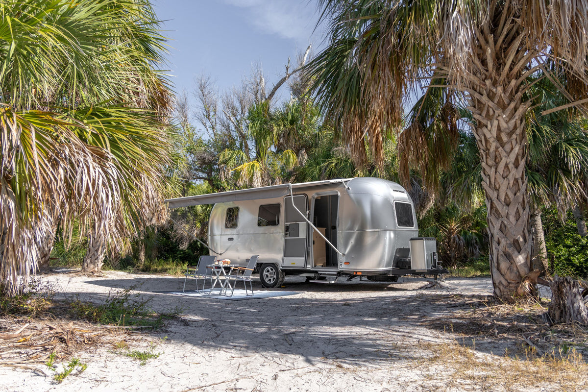A gleaming silver World Traveler travel trailer with an extended awning rests on sandy ground beneath palm trees, where a small table and chairs offer a shaded outdoor retreat. Nearby, the Airstream Cut Key Bundle for World Traveler Travel Trailers ensures peace of mind for any traveler enjoying this relaxing escape.