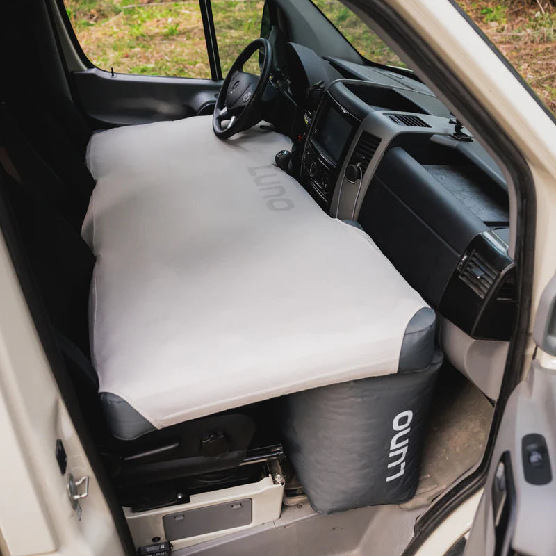 View of a van’s front cabin with a mattress covering both front seats. A gray cushion labeled Luno fills the footwell space, creating a flat sleeping surface from the driver’s to passenger’s side. For added mattress protection and comfort, using the Touring Coach Fitted Sheet for Luno Mattress is recommended.