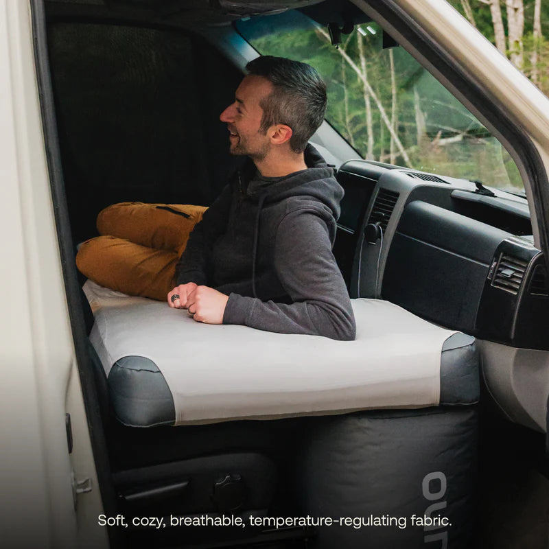 A person sits comfortably on a gray Luno air mattress in a van, smiling and gazing out the open door. Pillows rest beside them, with trees visible outside. Text reads: Soft, cozy, breathable, temperature-regulating fabric—Touring Coach Fitted Sheet for Luno Mattress included for enhanced comfort and mattress protection.