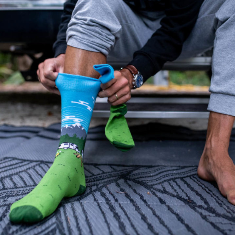A person sitting outdoors near an Airstream travel trailer puts on a bright green and blue pair of Airstream Campfire Socks featuring a mountain and tree design. One foot is bare, and the person, dressed in gray sweatpants and a watch, sits on a patterned mat.