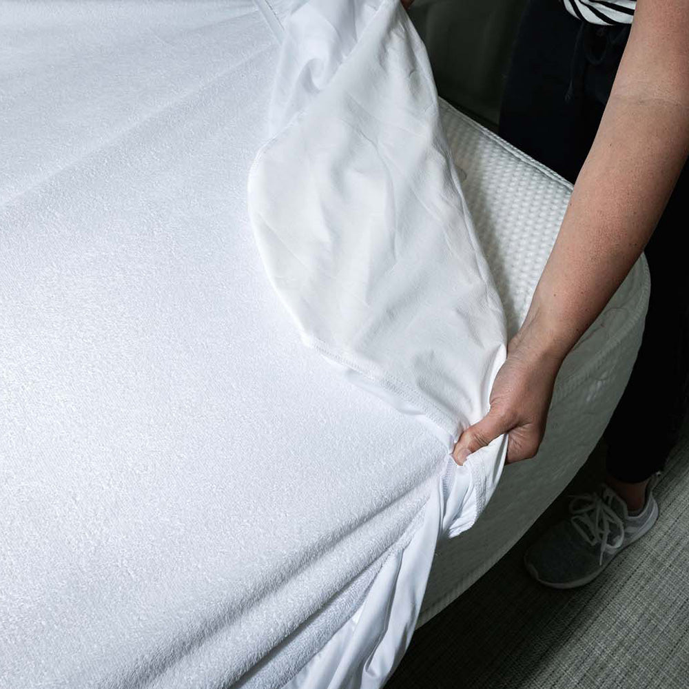 A person tucks an Airstream Custom Encasement for Classic Travel Trailers over a mattress, adjusting the corner. Only their hands and part of their arms are visible. The bed, protected by the custom encasement, is in a modern, well-lit room with neutral colors.