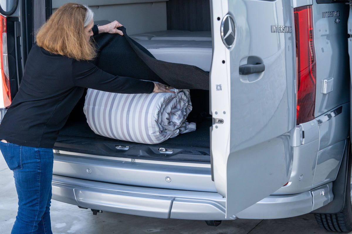 A woman lifts a black cover to access the back storage compartment of a van, placing a rolled-up blanket inside—perhaps preparing her Airstream x RVSuperbag All-In-One Bedding System for another comfortable night’s sleep on the road.