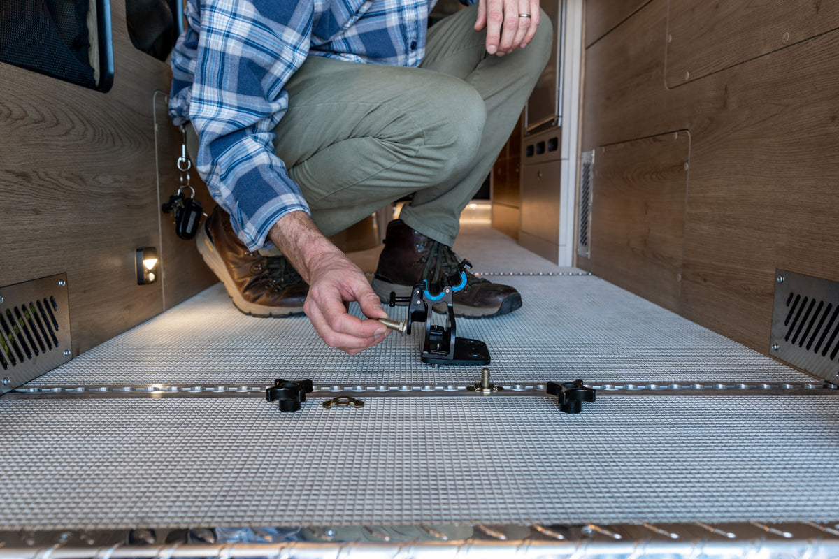 Wearing a plaid shirt and boots, a person kneels on the carpeted floor inside a wooden-paneled vehicle interior, installing an L-Track Bike Mount Bundle for Basecamp REI, Interstate 24X, Interstate 19X, and Rangeline by attaching the bike fork mount to the L-Track system.