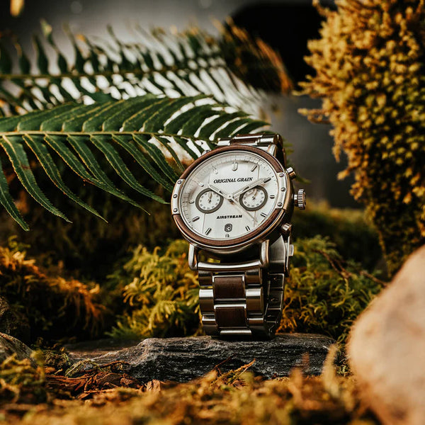 Silver watch with a detailed face on a wooden surface surrounded by green foliage