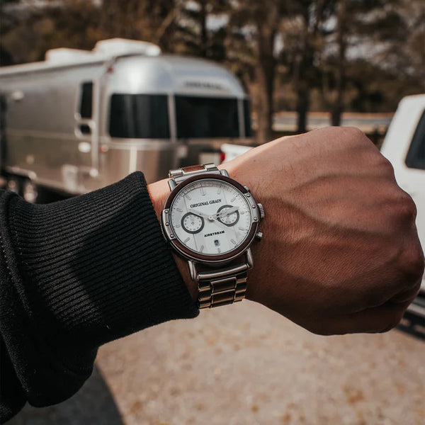Hand wearing a watch with a scenic background featuring a trailer and trees.