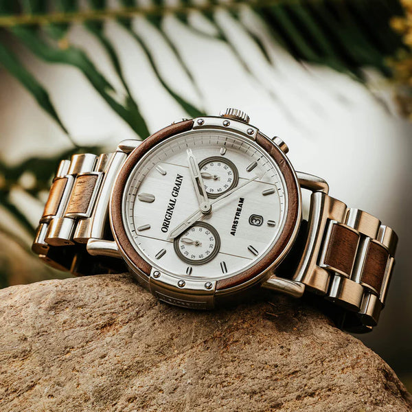 A wristwatch with a silver-tone metal and wood-tone bracelet, featuring a white dial with two subdials and a date window, resting on a textured stone with blurred green foliage in the background.