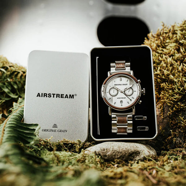 A wristwatch with a silver-tone metal bracelet and white dial sits in an open presentation box beside its silver lid, which reads AIRSTREAM ORIGINAL GRAIN. The box is styled on moss and rocks.