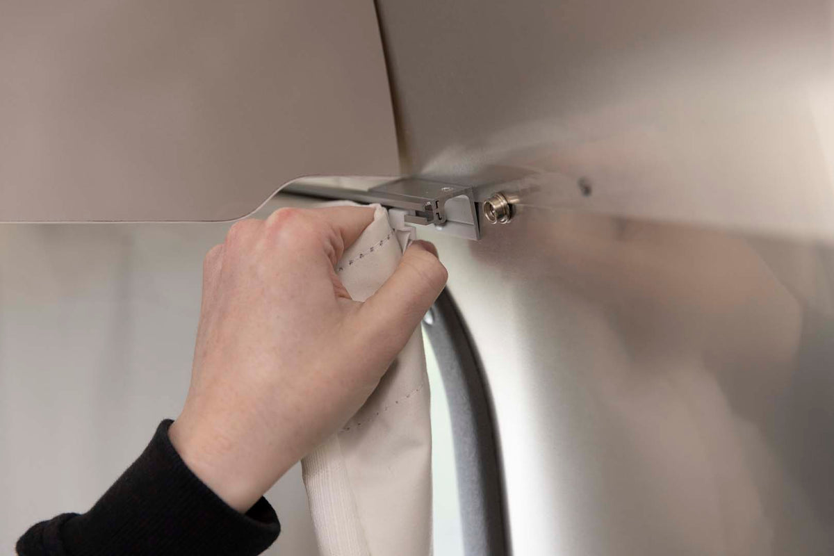 A persons hand attaches a white curtain to a metal hook on a curved, metallic wall, possibly inside a trailer or vehicle.